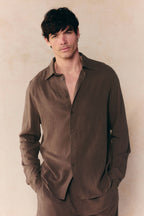Man in a relaxed-fit brown Cami Linen Classic Collar Shirt, unbuttoned at the top, hands in pockets.