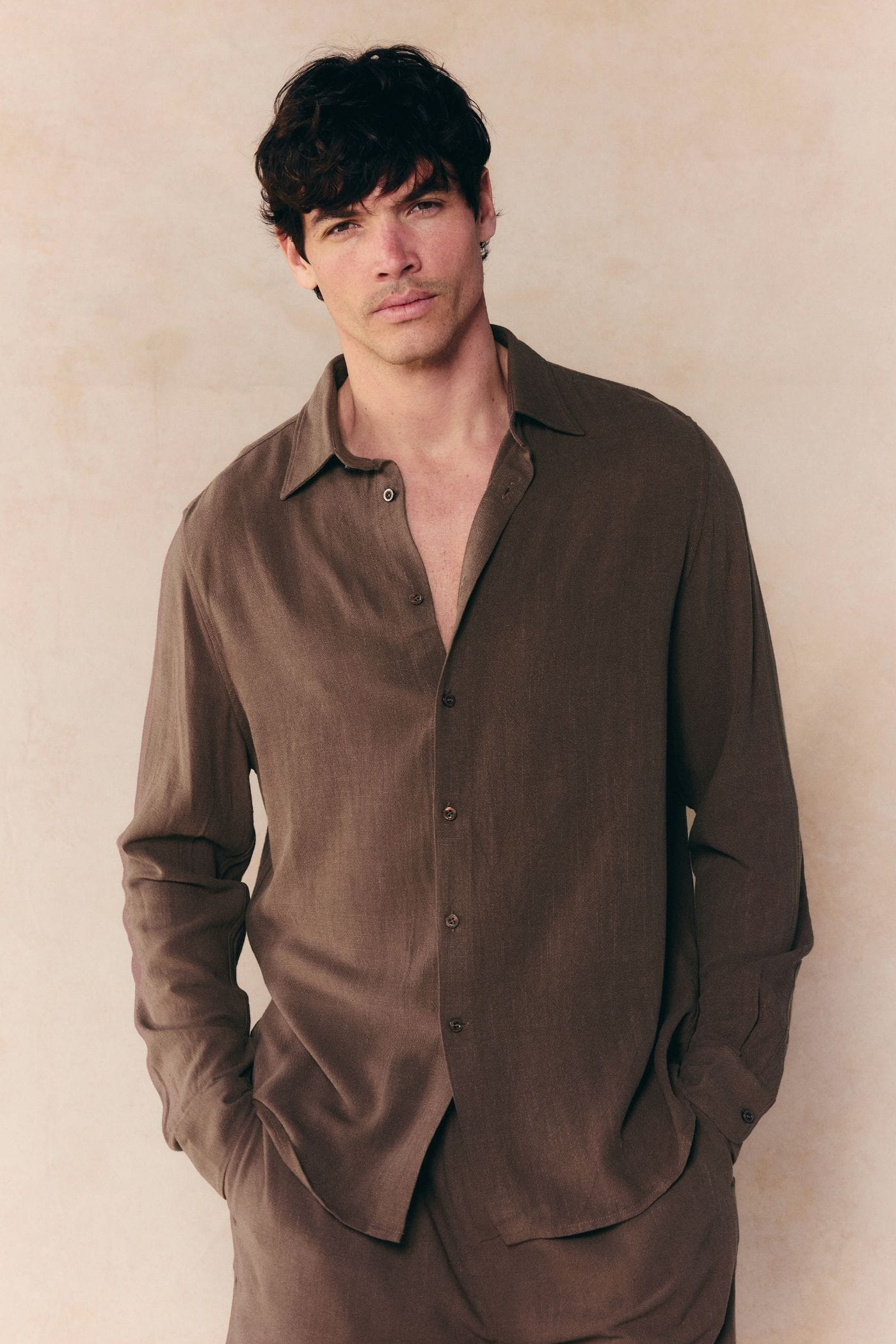 Man in a relaxed-fit brown Cami Linen Classic Collar Shirt, unbuttoned at the top, hands in pockets.