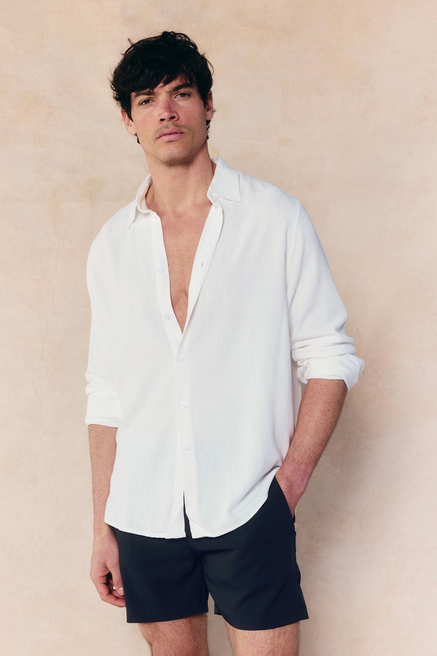 Man in an ecru Cami Linen Classic Collar Shirt, unbuttoned, with black shorts.