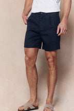 Men's navy linen pleated waistband shorts with side pockets, worn with a white t-shirt and sandals.