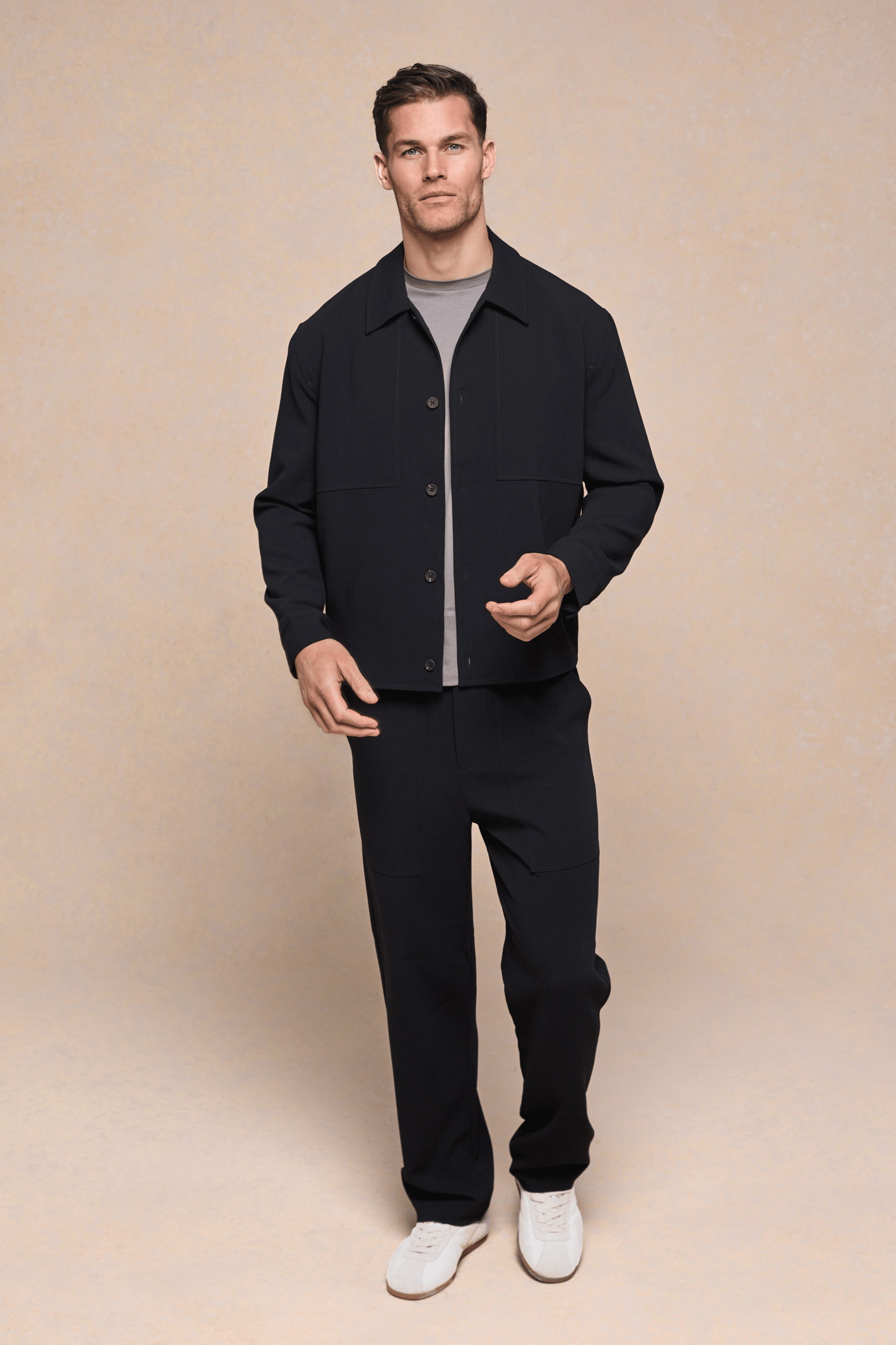 Man wearing black Julien Relaxed Panelled Trousers and matching jacket with a beige t-shirt and white sneakers.