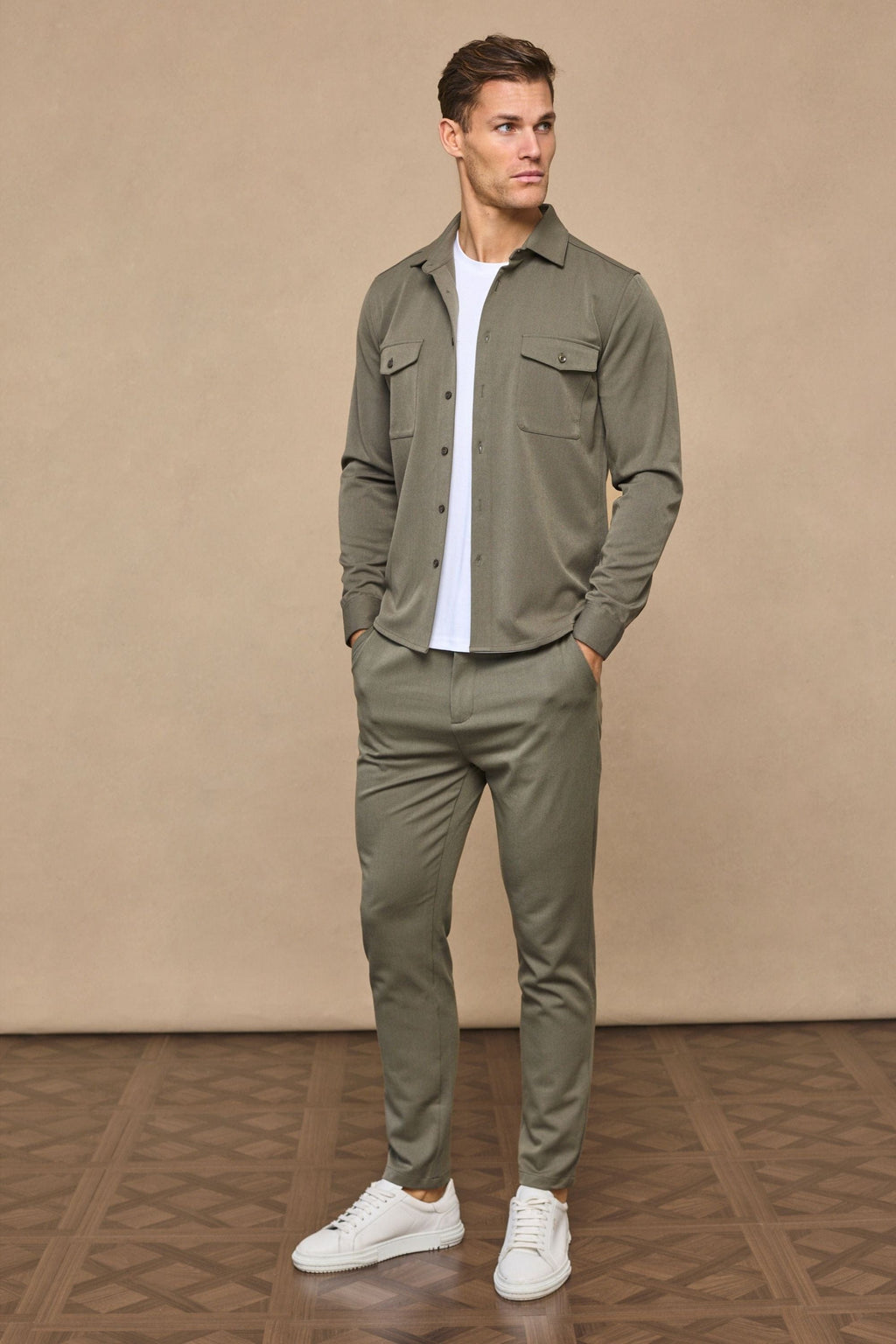 Man wearing a dark olive Leo overshirt with dual chest pockets, white t-shirt, matching trousers, and white sneakers.