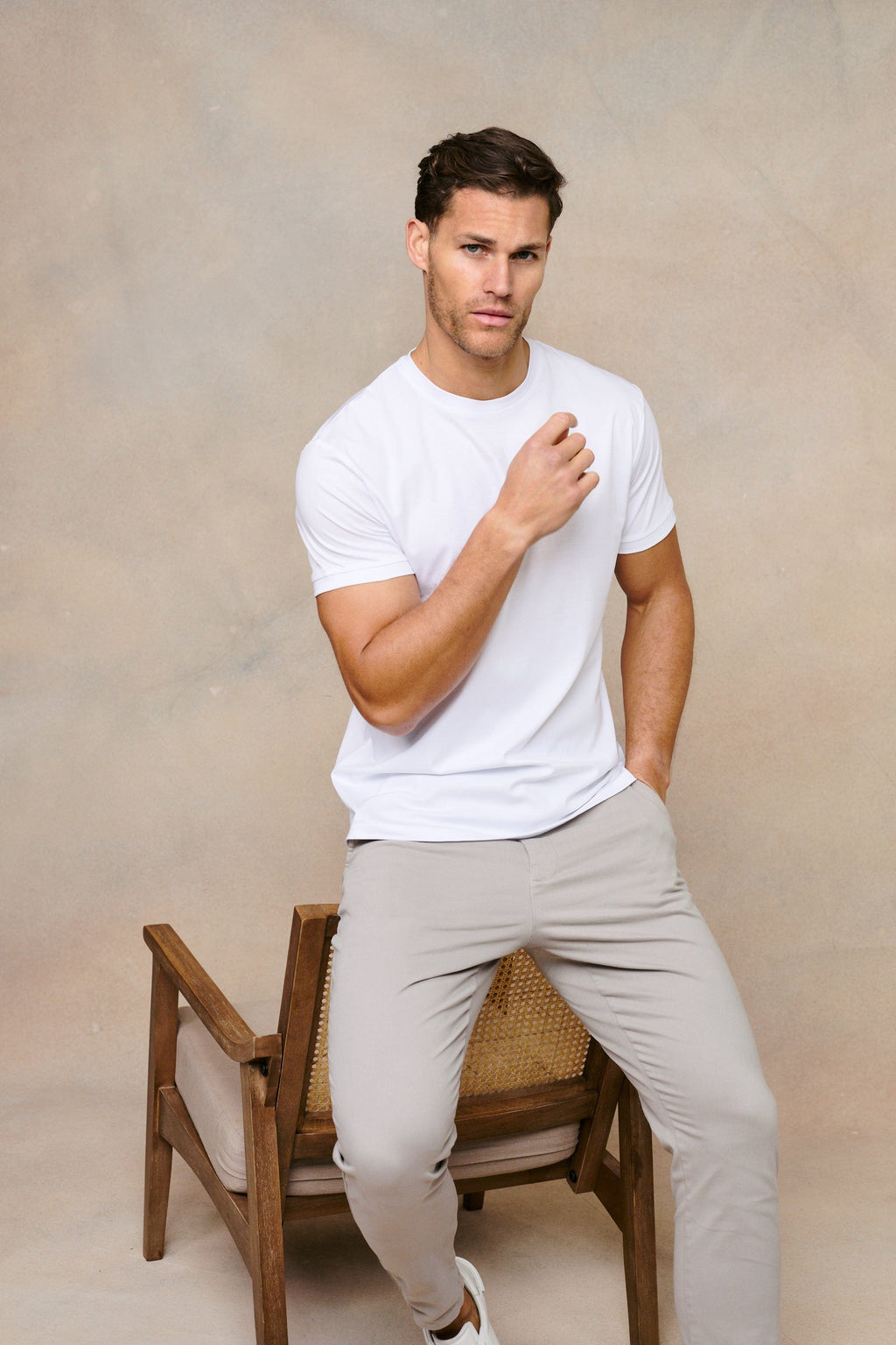 Man in a white mercerised cotton crew-neck t-shirt and light grey trousers, sitting on a wooden chair.