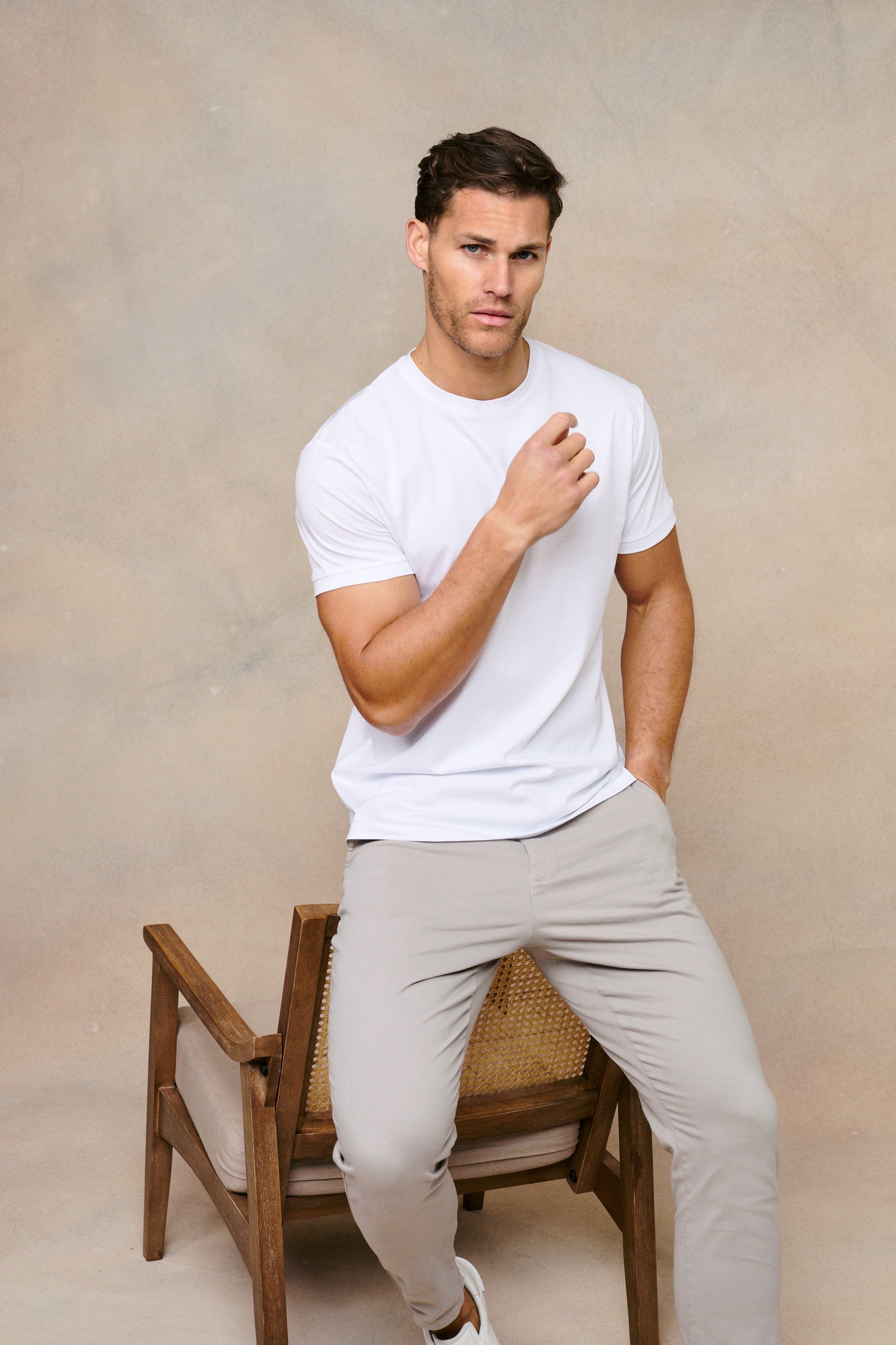Man in a white mercerised cotton crew-neck t-shirt and light grey trousers, sitting on a wooden chair.