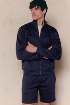 Man in a navy Luxe Zip Through Jacket and matching shorts, arms crossed, looking left.