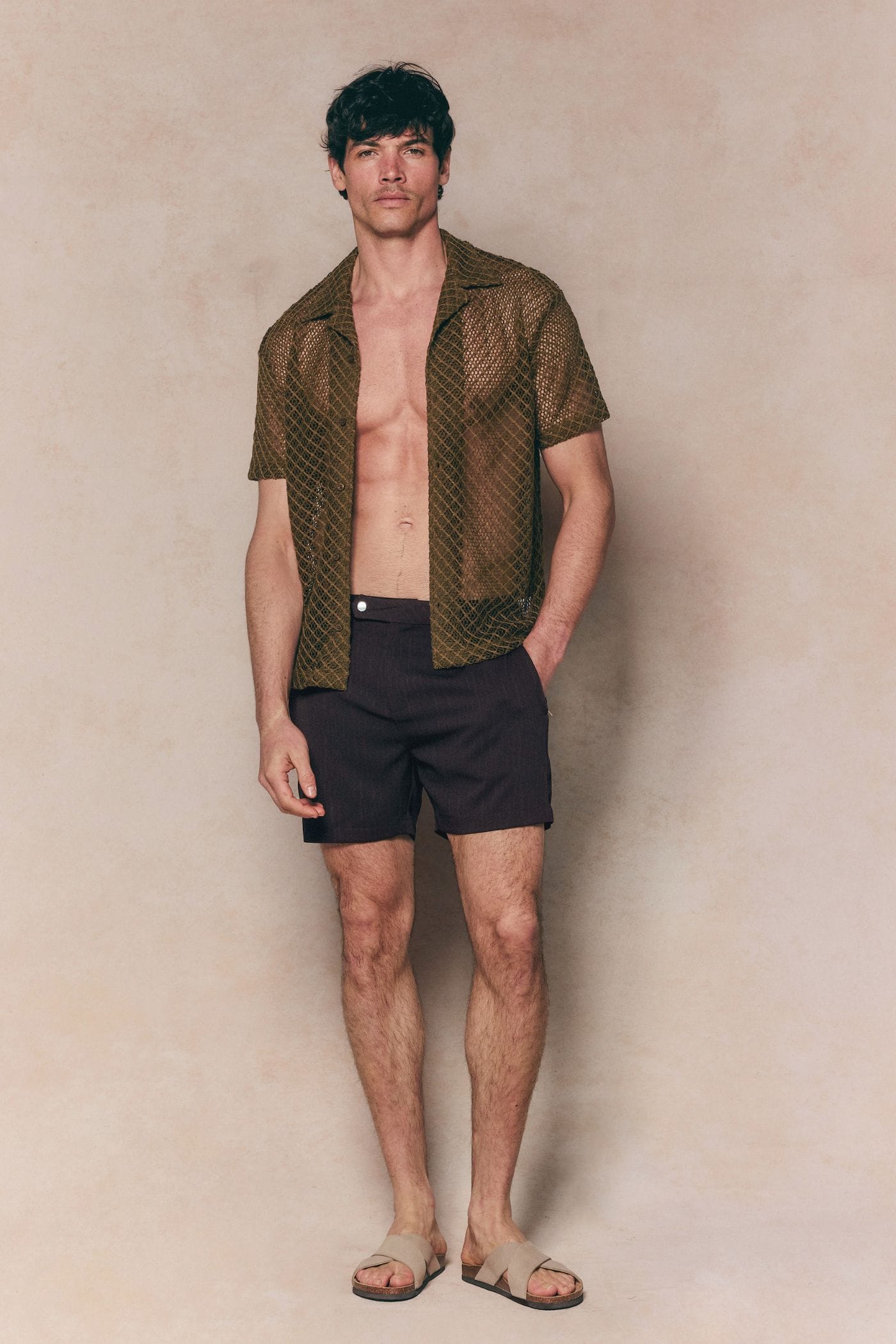Man wearing a brown Marcelo short sleeve lace shirt, unbuttoned, with dark shorts and sandals.