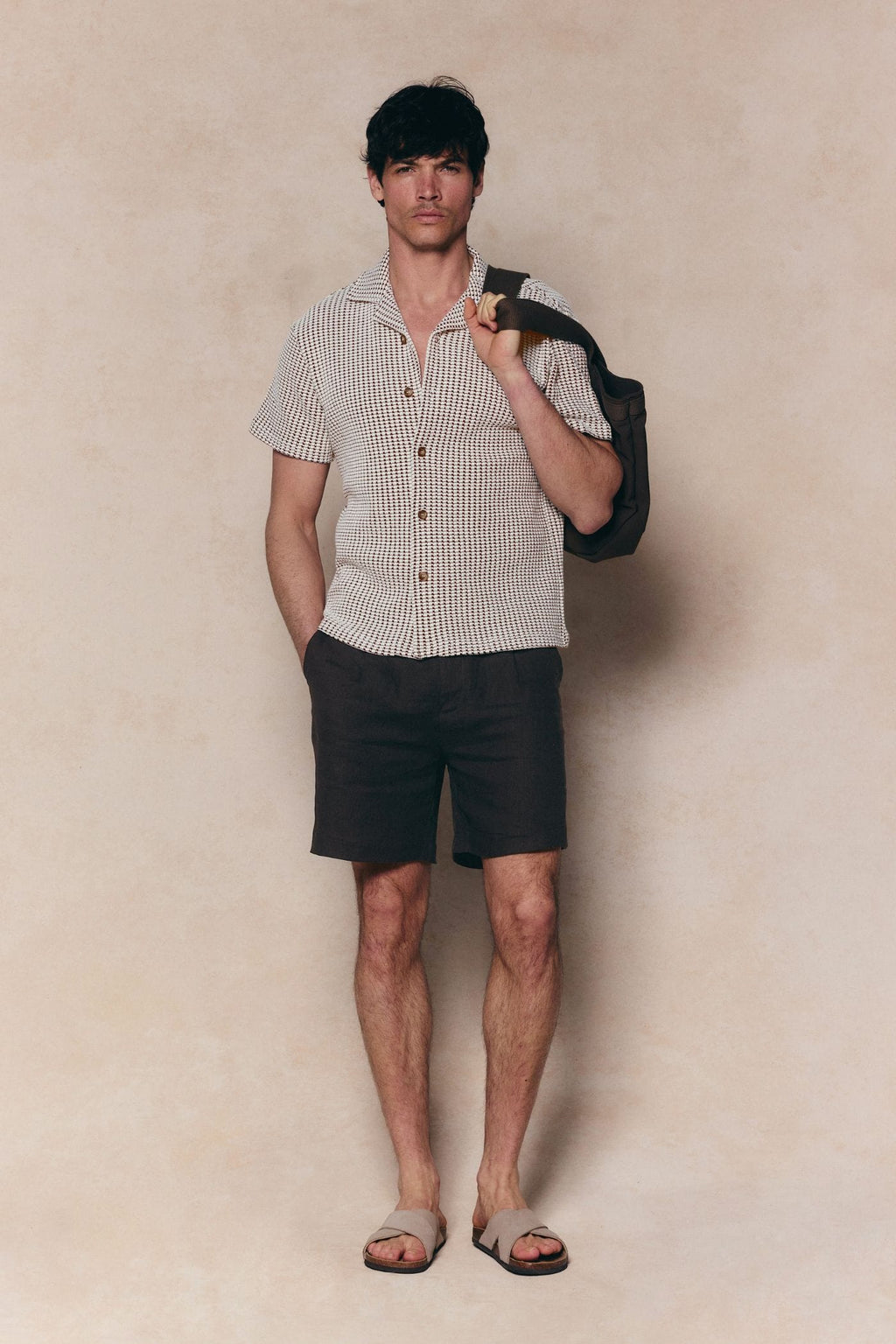 Man in Marcos white/brown patterned waffle short sleeve shirt, dark shorts, and sandals, holding a bag.