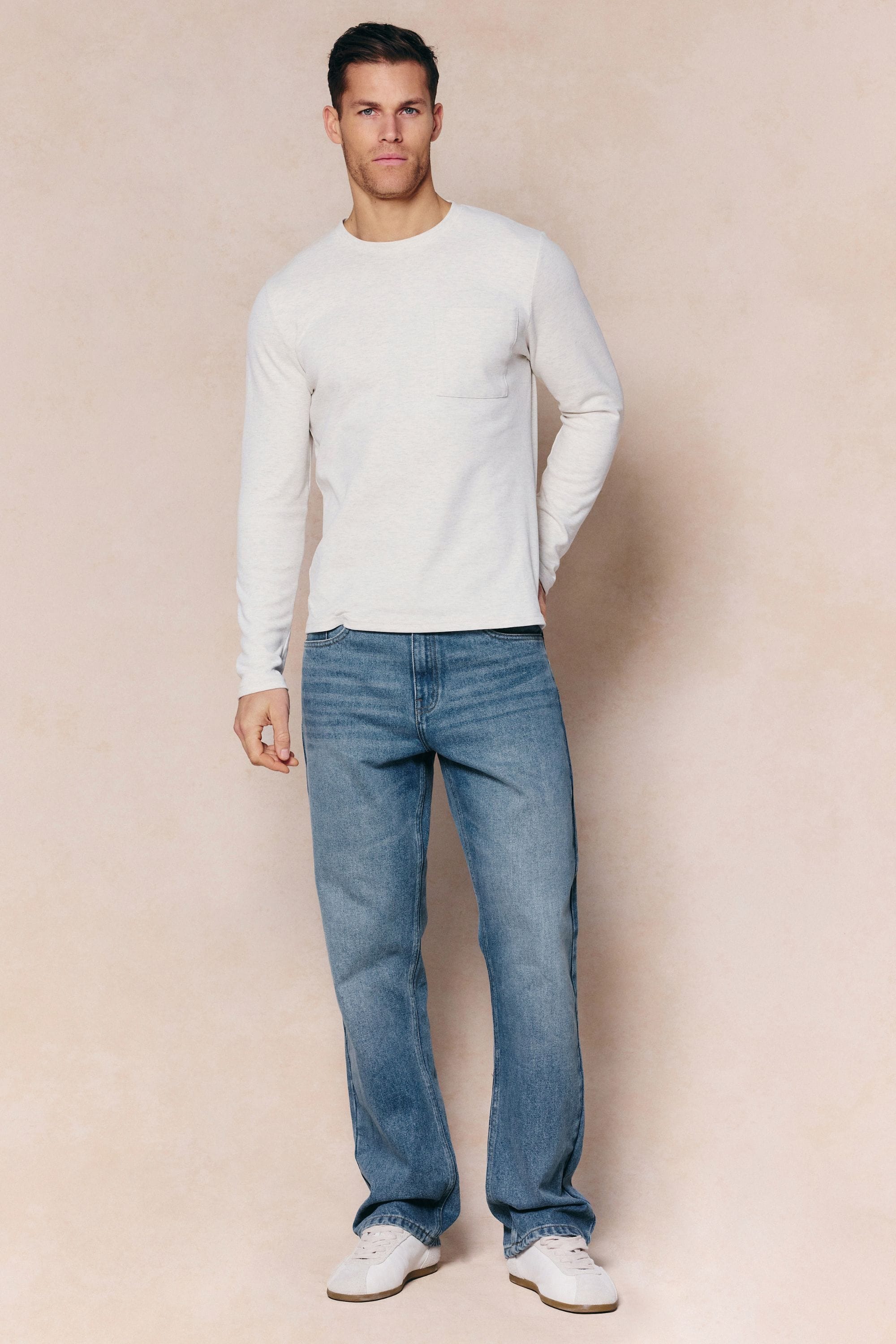 Man wearing Mason Relaxed Fit Midwash Indigo Jeans with a tan leather back patch and a cream long-sleeve shirt.