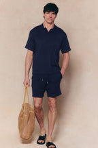 Man wearing a navy Miguel Boucle Polo Shirt and matching shorts, holding a woven tote bag.
