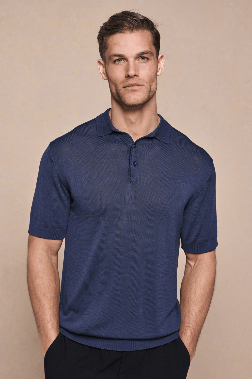 Man in a navy knitted short-sleeve polo with a half placket and ribbed sleeves, hands in pockets.