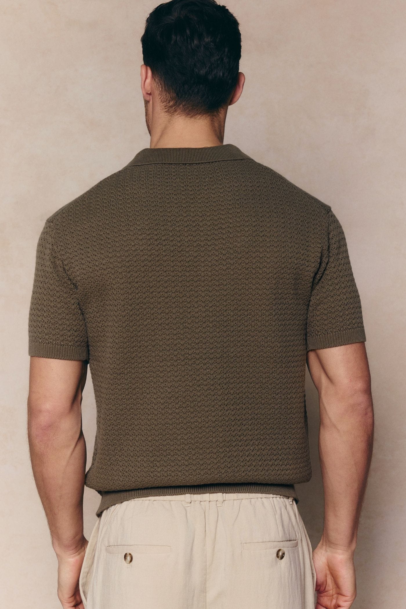 Man from behind wearing a dark olive Rapha cotton knit polo with textured finish and ribbed hem.