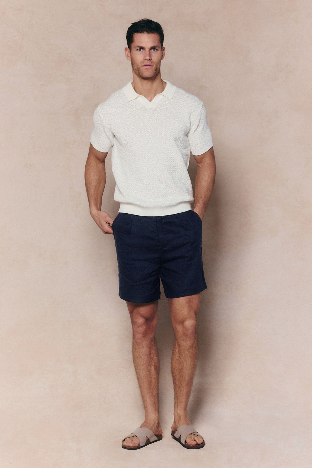 Man wearing an ecru Rapha cotton knit polo with open collar, navy shorts, and sandals.