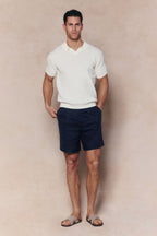 Man wearing an ecru Rapha cotton knit polo with open collar, navy shorts, and sandals.