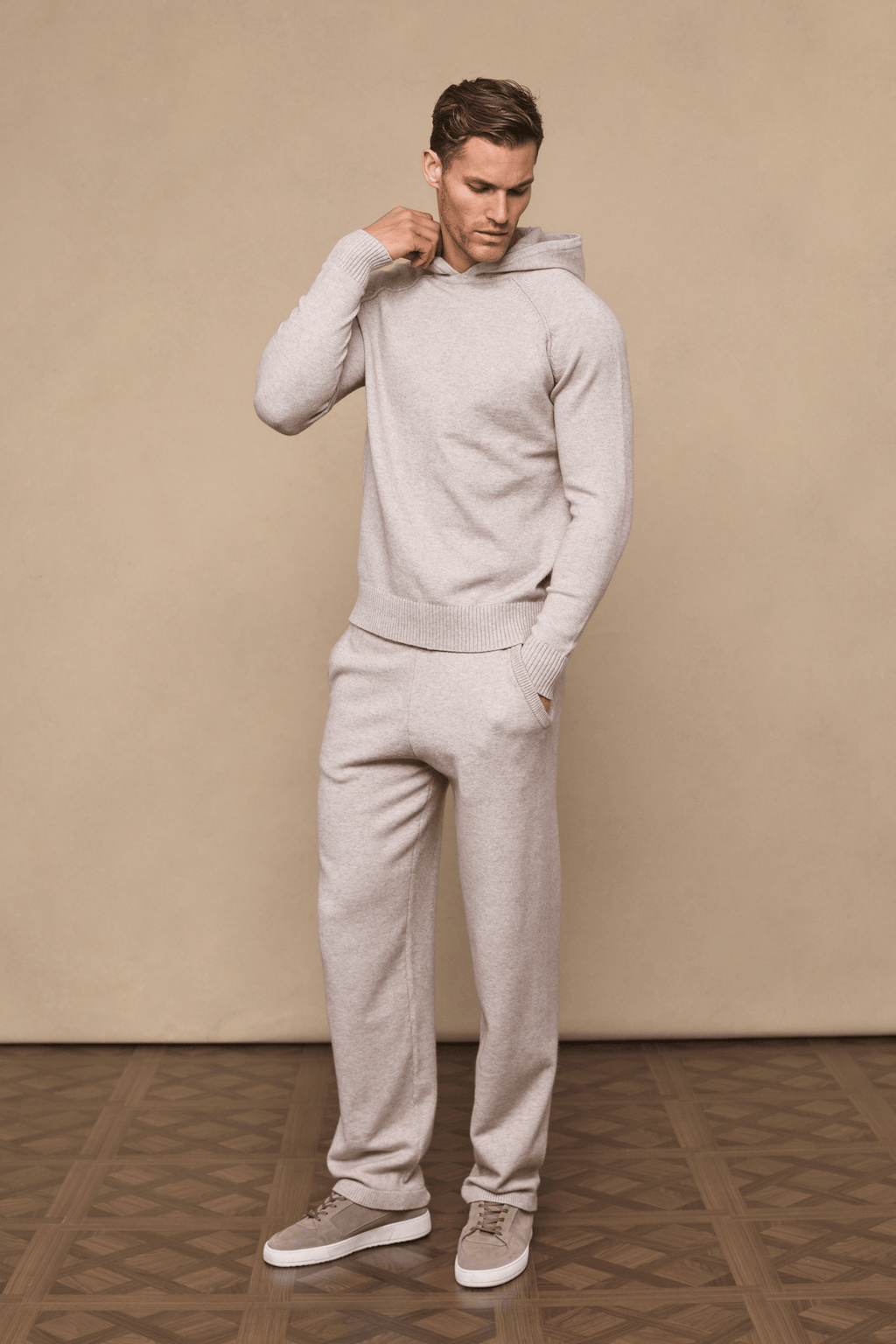 Man wearing an oatmeal Rowan Knitted Trouser and matching hoodie, hands in pockets, on a beige background.