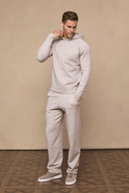 Man wearing an oatmeal Rowan Knitted Trouser and matching hoodie, hands in pockets, on a beige background.