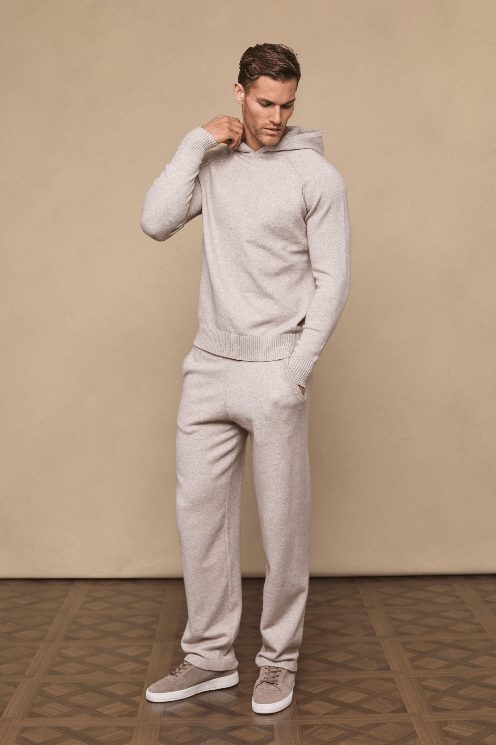 Man wearing an oatmeal Rowan Knitted Trouser and matching hoodie, hands in pockets, on a beige background.