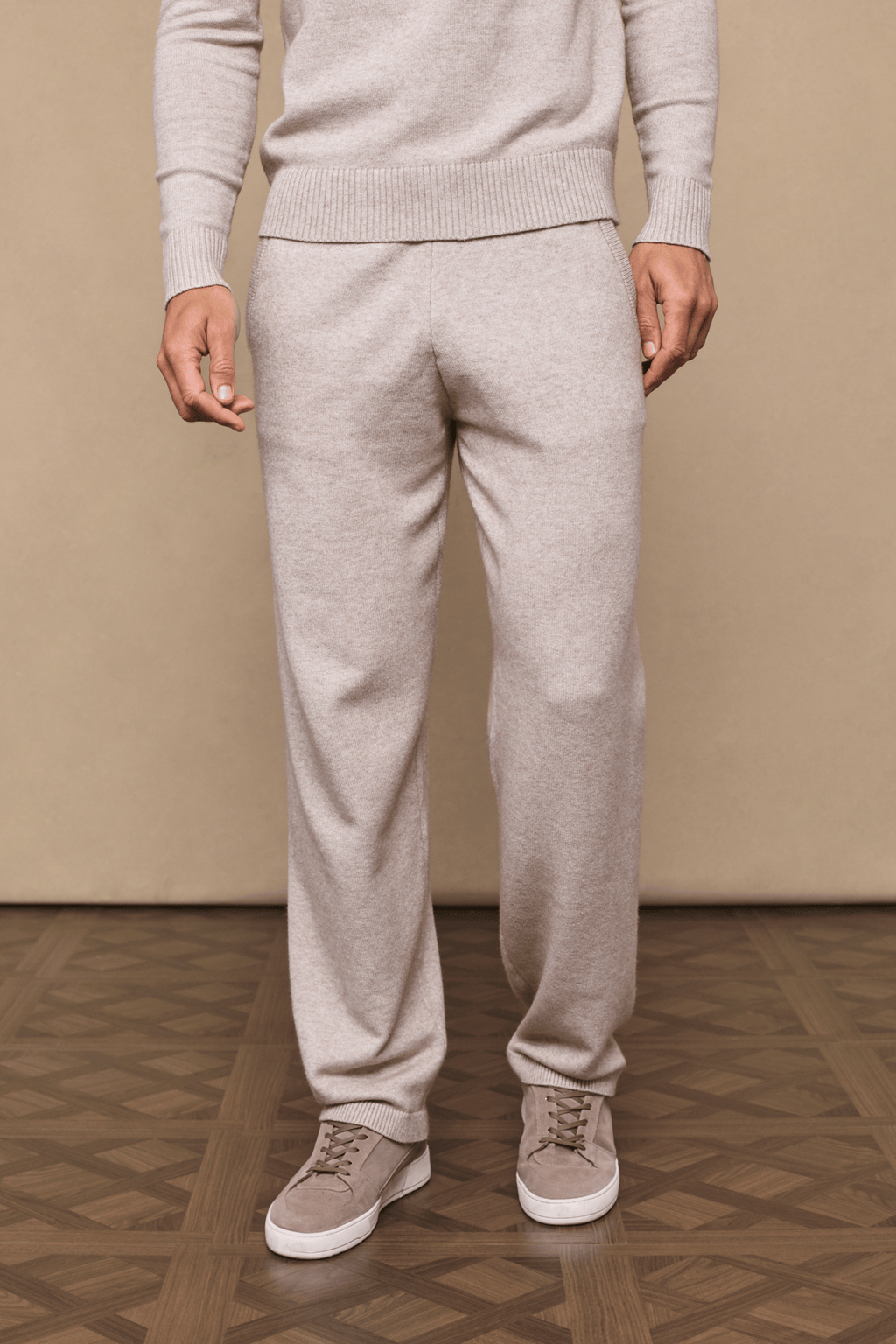 Man wearing oatmeal knitted trousers and matching sweater, with side pockets and drawcords, paired with beige sneakers.