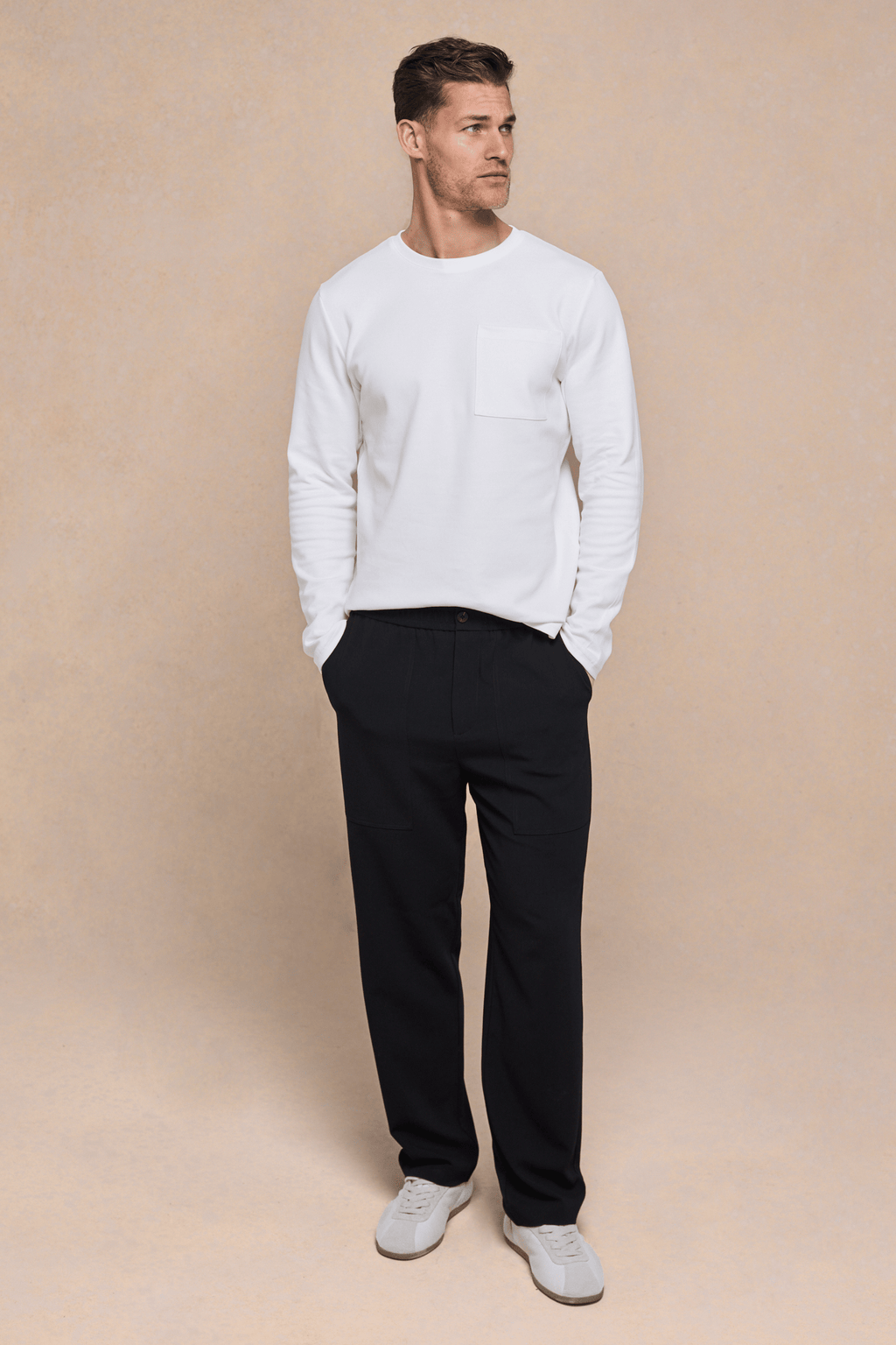 Man in ecru long sleeve crew neck t-shirt with chest pocket, black pants, and white sneakers.